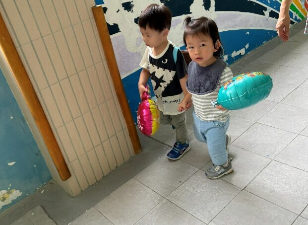 聽損幼兒在融合體驗活動中與同學手牽手走在走廊上，每人各拿一個氣球，展現聽常孩子的理解接納與同儕情誼。